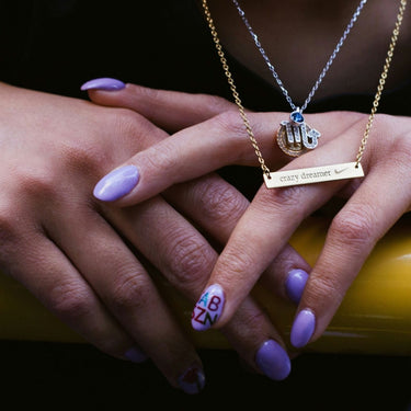 gold bar necklace and hamsa pendant necklace on hands with purple manicured nails