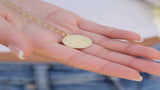 A hand holding a gold stainless steel charm necklace with a LOVE pendant, shown in natural daylight for detail.